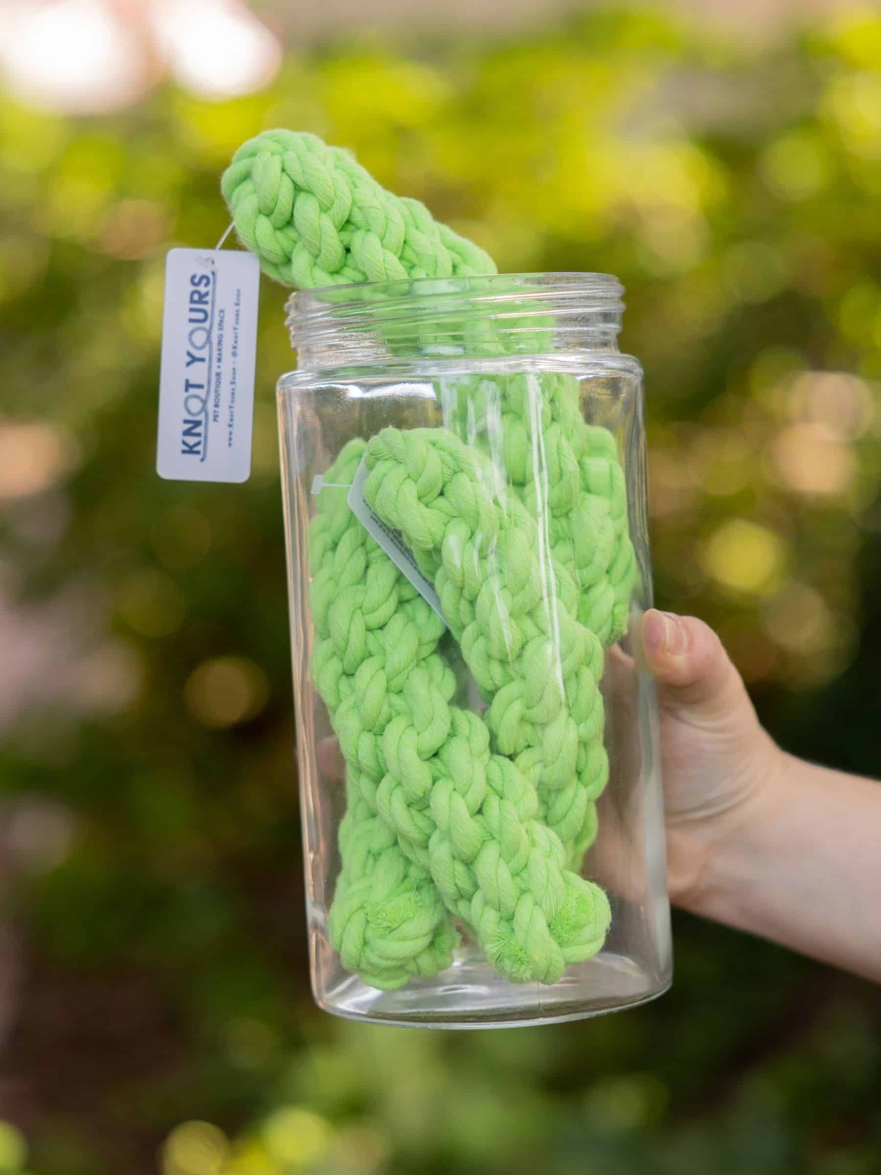 A large clear jar full of green knotted rope toys shaped like pickles held by a hand with a blurred green background