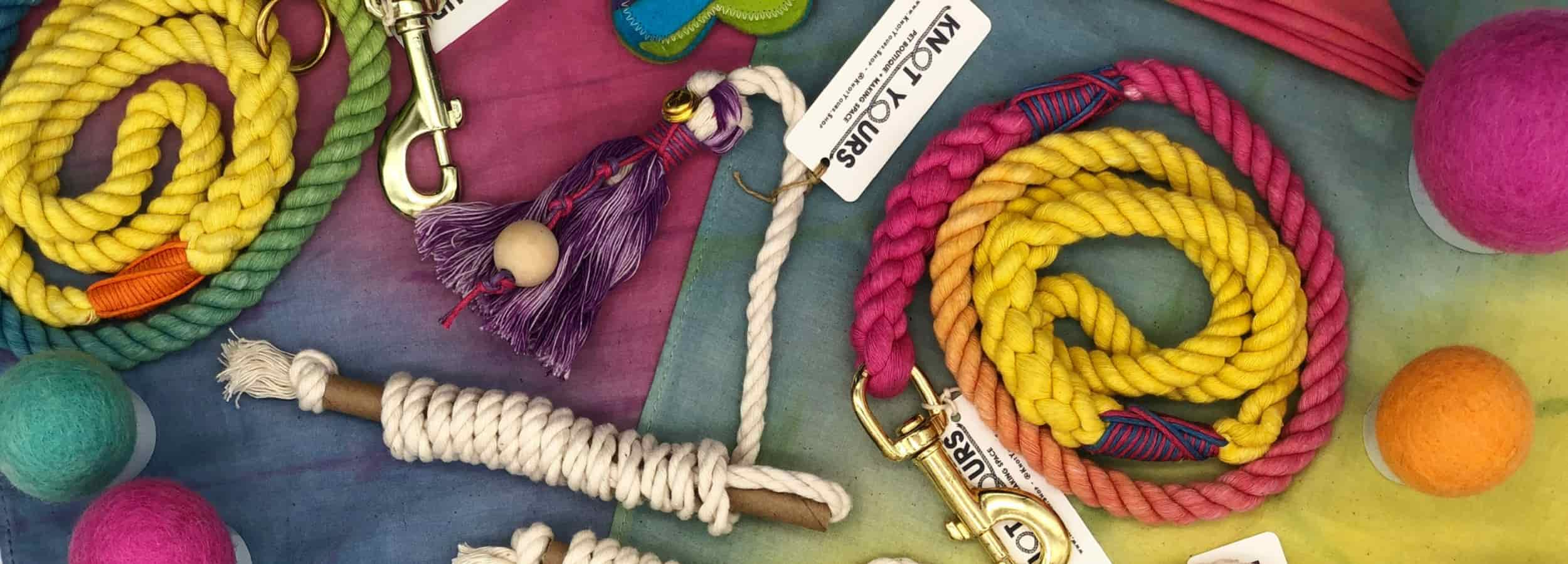 A vibrant flat lay of handmade dog accessories from Knot Yours, including colorful rope leashes, tug toys, felt balls, and branded tags, all arranged on a rainbow-dyed fabric background.