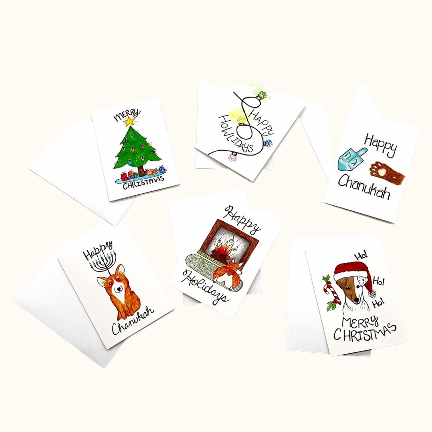 Six illustrated mini holiday cards laid out with white envelopes, featuring cheerful drawings like a Christmas tree, dreidel, menorah, and cozy fireplace scene.