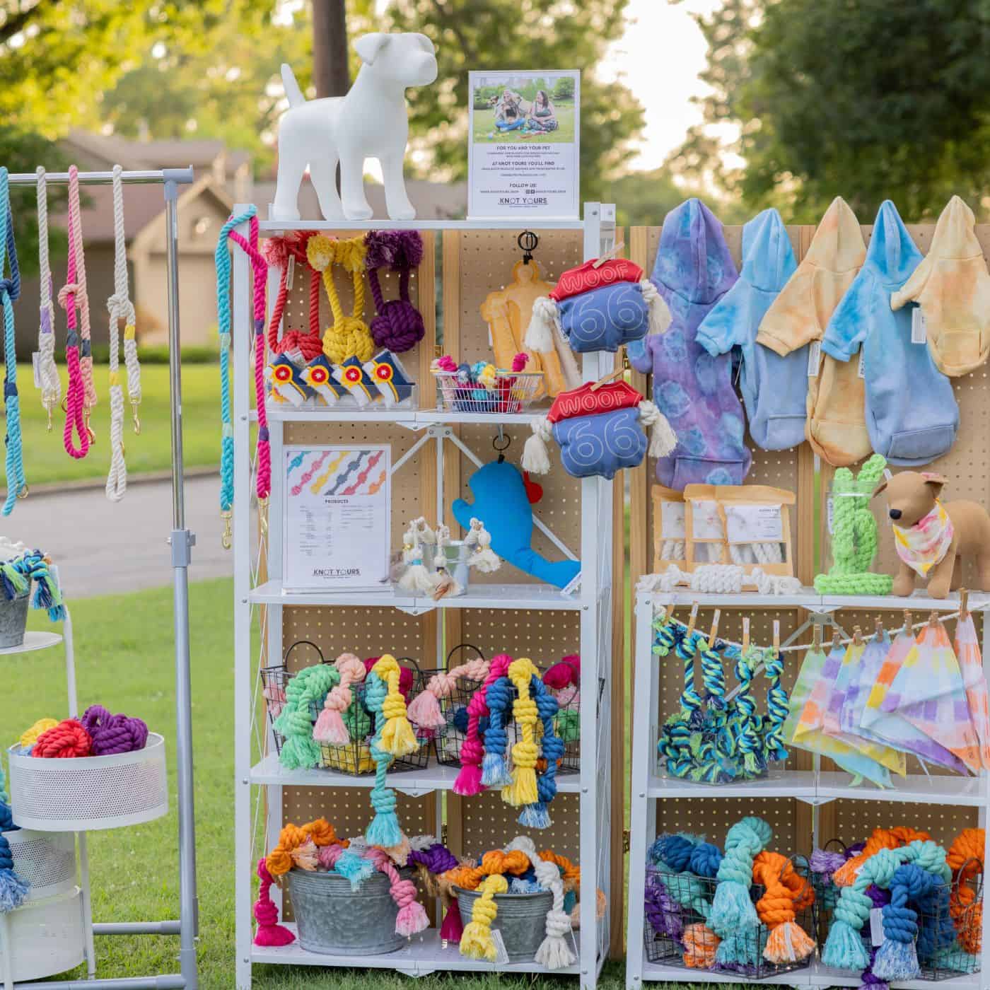 Outdoor market display of colorful rope tug toys, plush toys, tie-dye dog hoodies, and dog bandanas on white shelves.