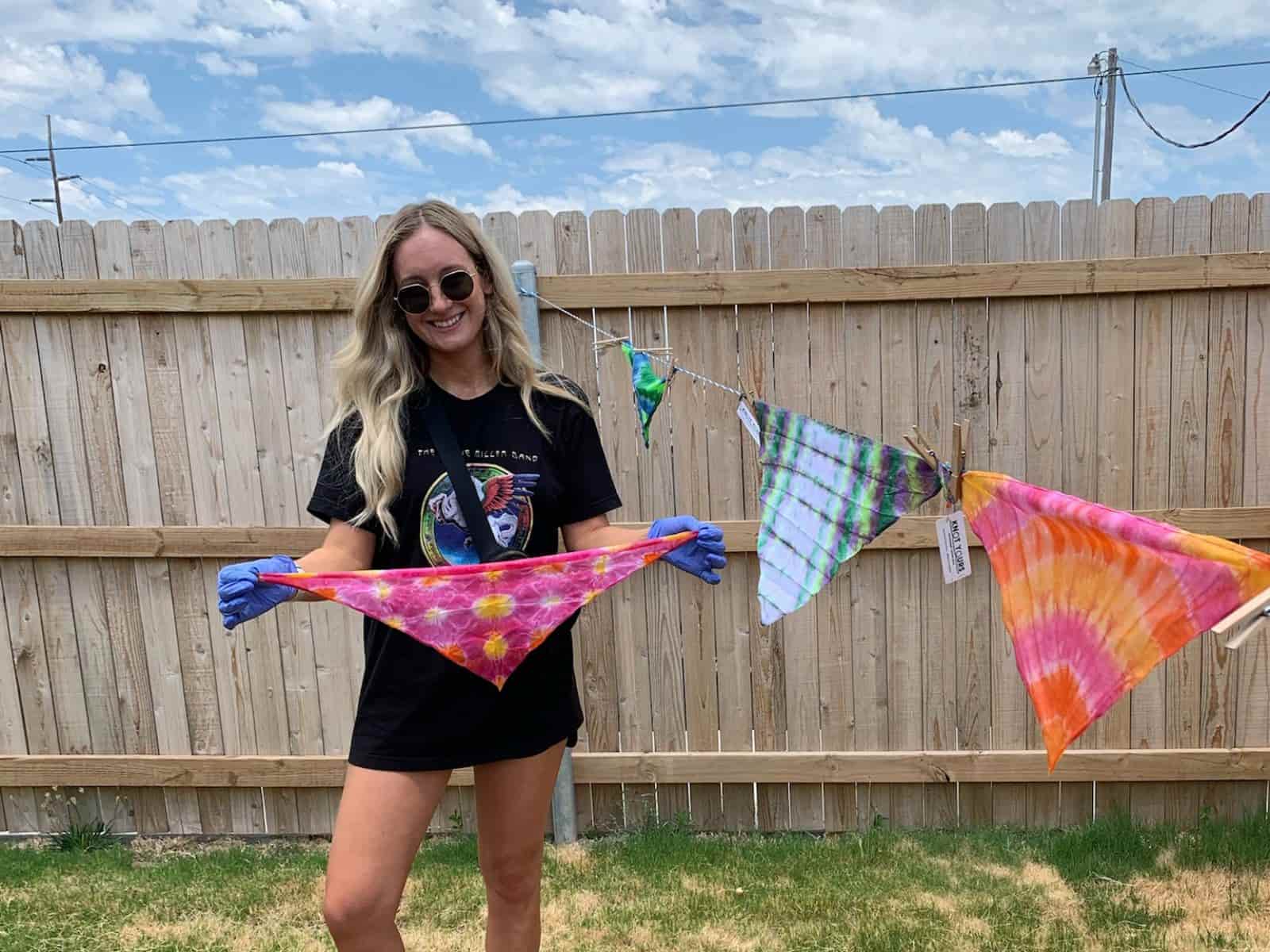 Woman holding tie-dye dog bandana in a backyard with a wooden fence and clothesline in the background.