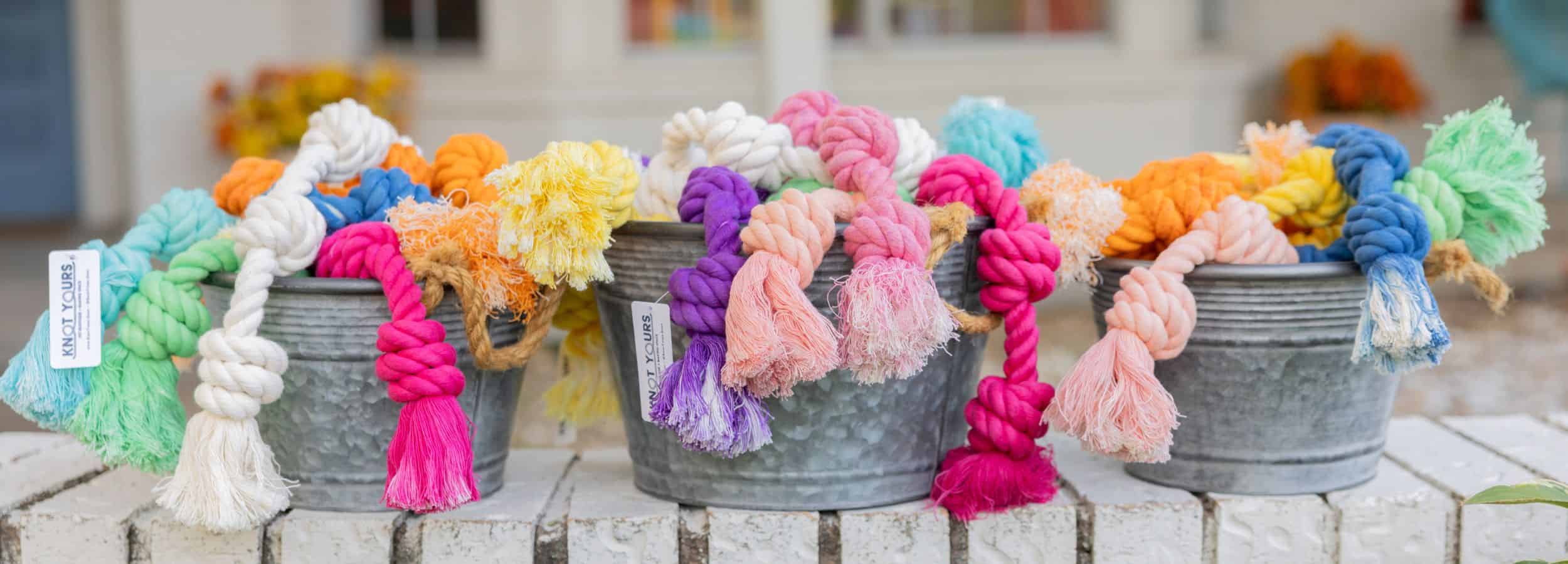 Colorful dog toys with rope handles in metal buckets on a wooden surface.