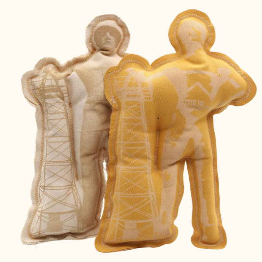 Two plush squeaky toys shaped like Tulsa’s Golden Driller statue, one in mustard yellow and the other in natural canvas, each screen-printed with oil rig tower details.