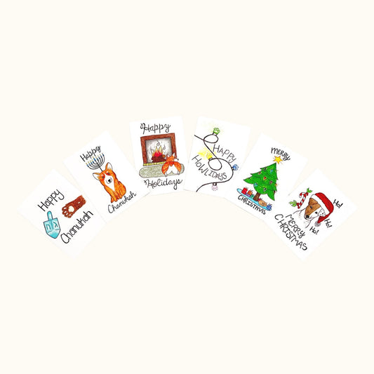 A fan display of six illustrated mini holiday cards with festive designs including menorahs, Christmas trees, a fireplace, and pets in holiday attire.