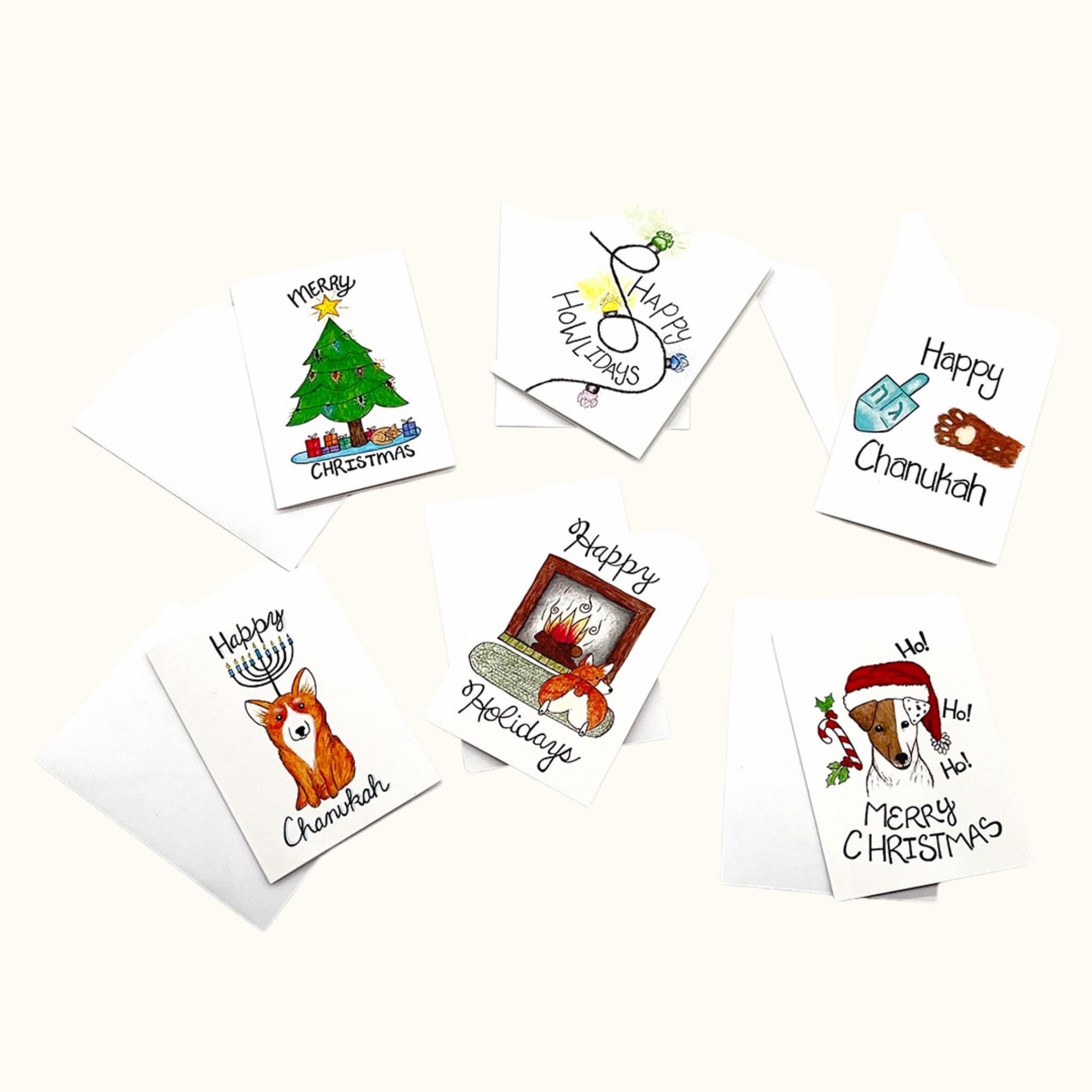 Six illustrated mini holiday cards laid out with white envelopes, featuring cheerful drawings like a Christmas tree, dreidel, menorah, and cozy fireplace scene.