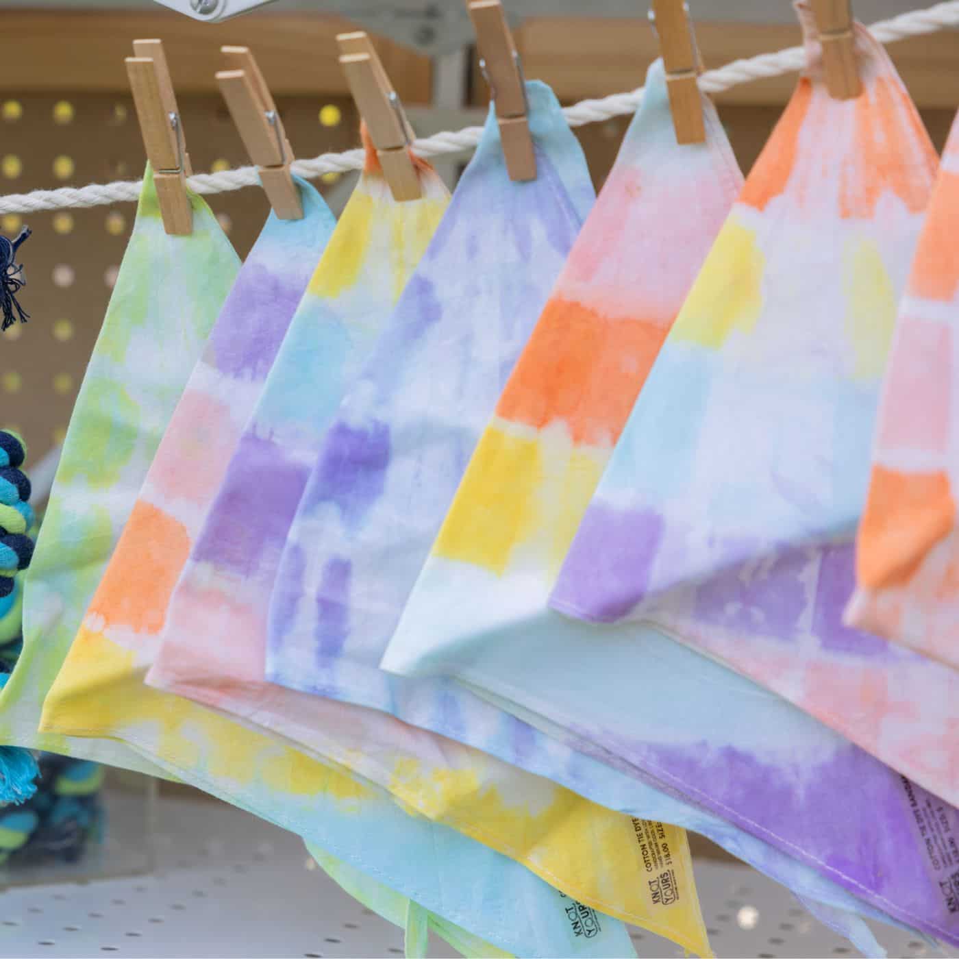 Pastel tie-dye dog bandanas in purple, yellow, orange, green, and blue hanging from a rope with wooden clothespins.
