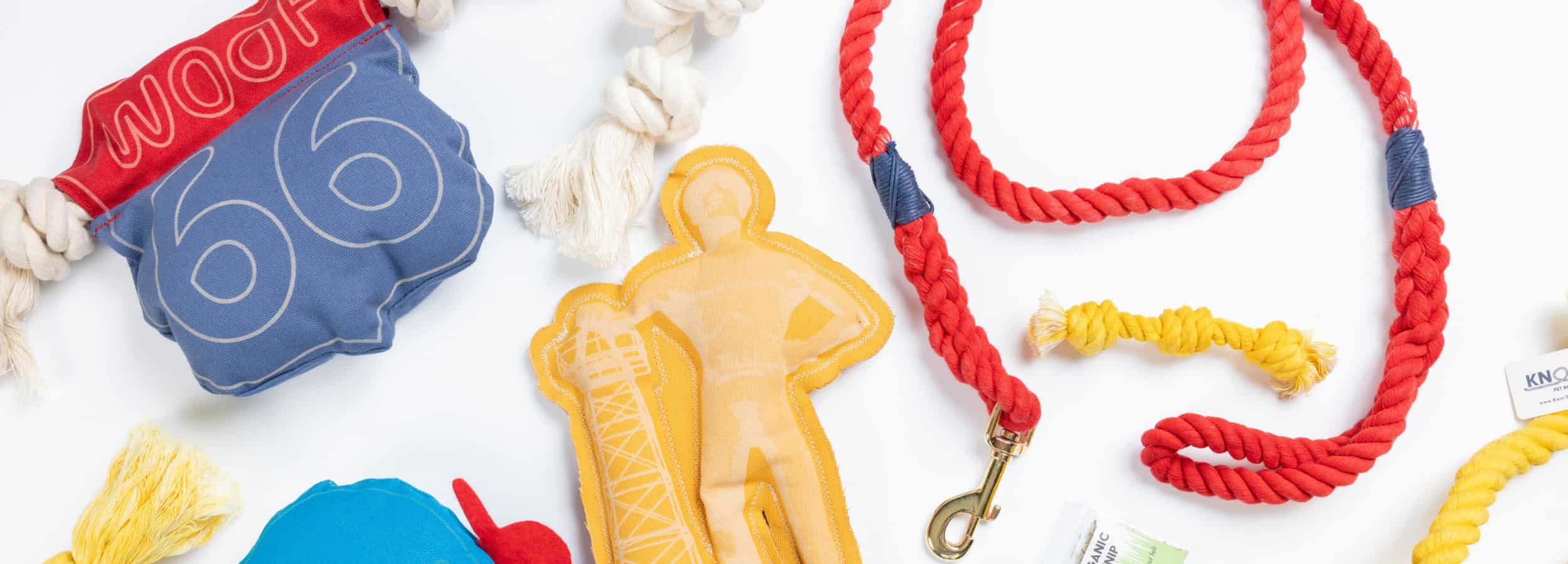Collection of dog toys made in the USA including a yellow fetch toy, woof 66-plush toy, a rope toy, and a lead on a white background.